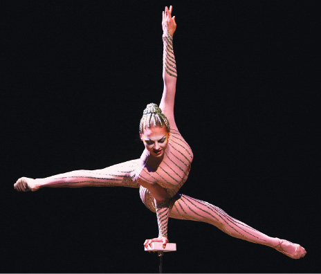 Imagem: Outra bailarina está com a mão direita apoiada em um suporte, a mão esquerda para cima e as pernas abertas.  Fim da imagem.