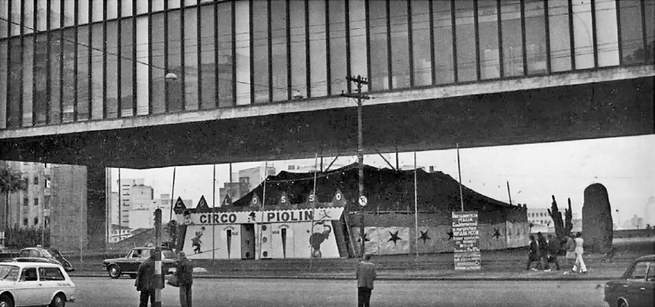 Imagem: Fotografia em preto e branco. Uma tenda grande de circo com uma placa e a informação: CIRCO PIOLIN. Em volta há algumas pessoas e ao lado, um carro em uma avenida. Acima do circo há uma construção extensa e retangular. Fim da imagem.