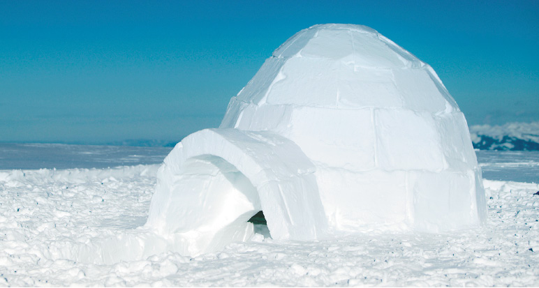 Imagem: Fotografia. Um iglu redondo feito de blocos de gelo. Em volta há muita neve e ao fundo, o céu azul. Fim da imagem.