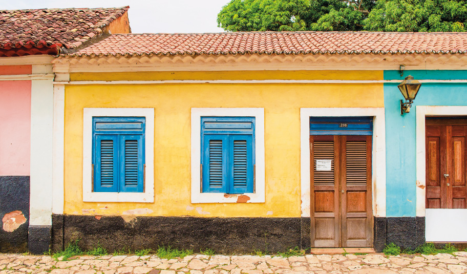 Imagem: Fotografia. No centro, uma casa térrea com a parede amarela na parte superior e preta na parte inferior. Duas janelas quadradas e azuis uma porta retangular marrom.  Fim da imagem.