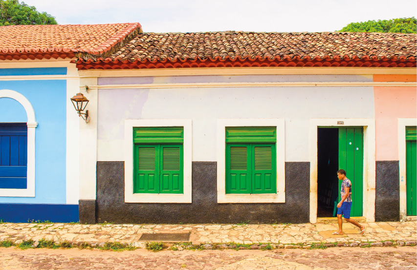 Imagem: Fotografia. No centro, uma casa térrea com a parede branca na parte superior e preta na parte inferior. Duas janelas quadradas e uma porta retangular verdes.  Fim da imagem.