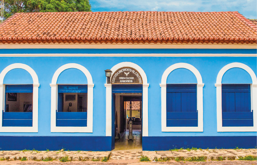 Imagem: Fotografia. Casa térrea com parede azul-claro, janelas retangulares azul-escuro com bordas arredondadas e brancas.  Fim da imagem.