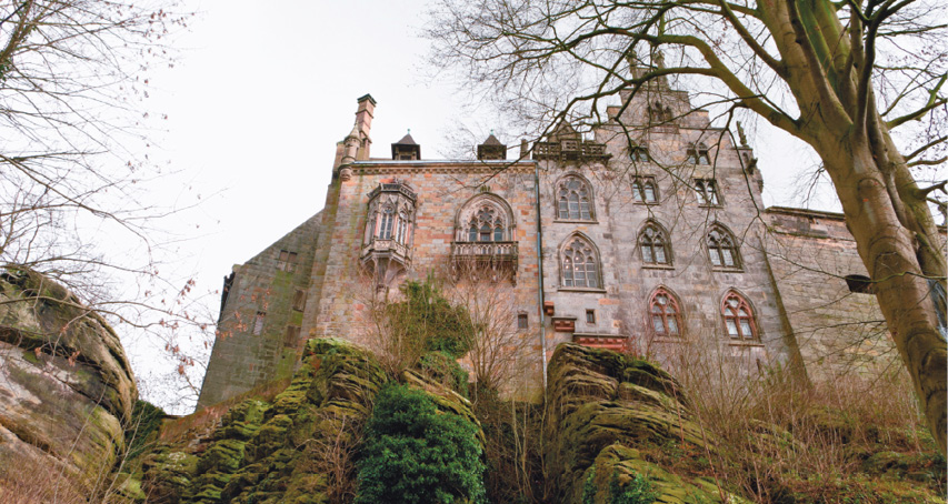 Imagem: Fotografia. Vista de baixo de um castelo grande com janelas arredondadas e árvores em volta. Fim da imagem.