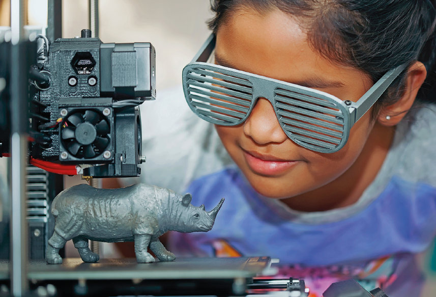 Imagem: Fotografia. Uma menina com cabelo preso e óculos cinza no rosto está sorrindo e observando uma impressora 3D, que está imprimindo um boneco cinza com formato de rinoceronte. Fim da imagem.