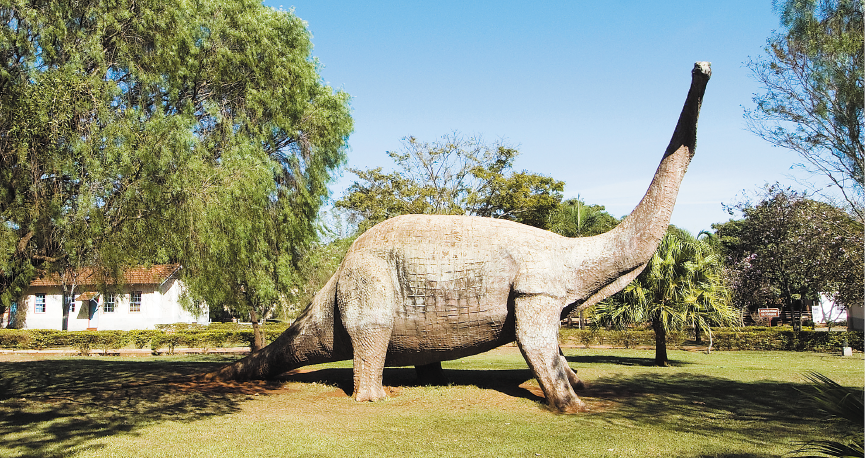 Imagem: Fotografia. Réplica de um dinossauro com pescoço e rabo compridos. Em volta há muitas árvores e ao fundo, uma casa.   Fim da imagem.