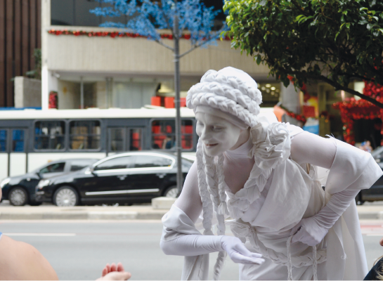 Imagem: Fotografia. Uma pessoa com o corpo todo pintado de branco está inclinada para frente com a mão direita virada para cima. Ao fundo, carros em uma avenida.   Fim da imagem.