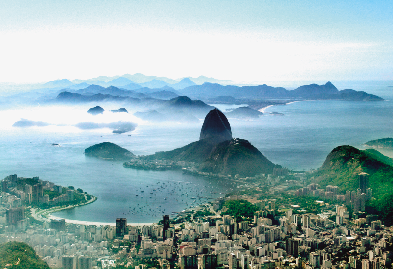 Imagem: Fotografia. Vista aérea de uma cidade ao lado do mar e ao fundo, morros. Fim da imagem.