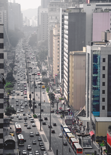 Imagem: Fotografia. No centro há automóveis enfileirados em uma avenida. Nas laterais há prédios e postes.   Fim da imagem.