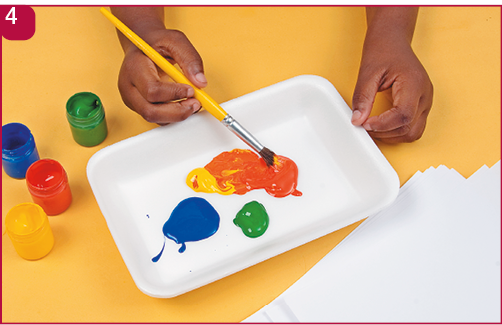 Imagem: Fotografia 4. Mão segurando o pincel sobre tintas em outra bandeja. Ao lado, potes de tinta e papéis em branco.  Fim da imagem.