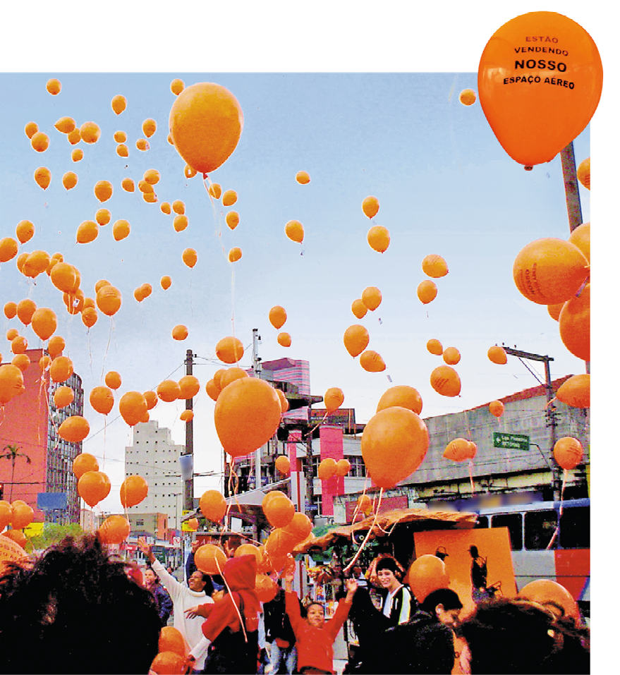 Imagem: Fotografia. Vários balões laranja voando. Abaixo há pessoas e prédios. No canto superior direito, destaque para um balão laranja com a informação: ESTÃO VENDENDO NOSSO ESPAÇO AÉREO. Fim da imagem.