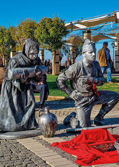 Imagem: Fotografia. Escultura de um soldado ajoelhado com a mão esquerda no peito e a direita na cintura. Atrás dele, escultura de uma mulher com vestido e lenço na cabeça. Ela está com o corpo inclinado para frente e segurando um pedaço de madeira com as duas mãos. Entre eles há um jarro no chão e na frente deles, um tecido vermelho.   Fim da imagem.