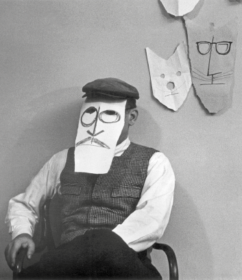 Imagem: Fotografia em preto e branco. Um homem com boina, colete, camisa e calça está sentado com uma máscara de papel na frente do rosto. Atrás dele há máscaras de papel penduradas na parede.  Fim da imagem.