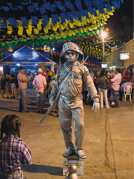 Imagem: Fotografia. Uma pessoa com roupa de cangaceiro está com o corpo todo pintado de prata e parado sobre um suporte. Na frente, uma menina observa e ao fundo há pessoas, barracas e bandeiras coloridas penduradas.  Fim da imagem.