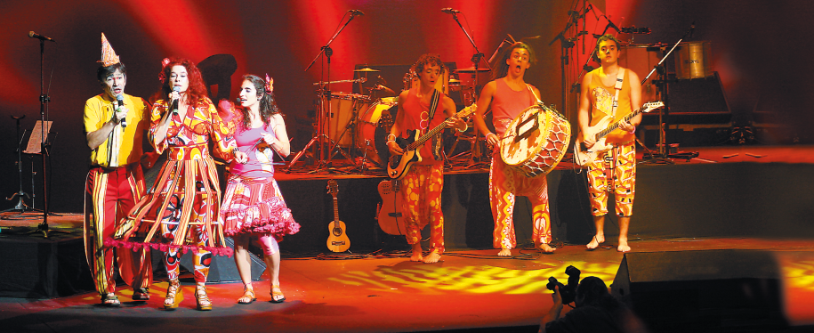 Imagem: Fotografia. À esquerda, duas mulheres com vestidos coloridos e um homem com chapéu pontudo estão cantando. À direita, três homens estão tocando instrumentos musicais. Ao fundo, mais instrumentos musicais.    Fim da imagem.