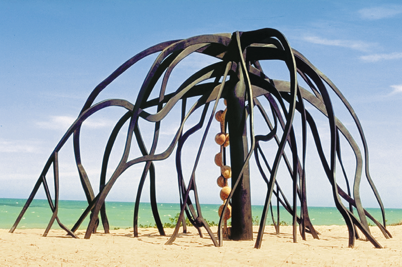 Imagem: Fotografia. Escultura com formato de tronco de árvore e com galhos compridos até o chão. Ao fundo, o mar. Fim da imagem.