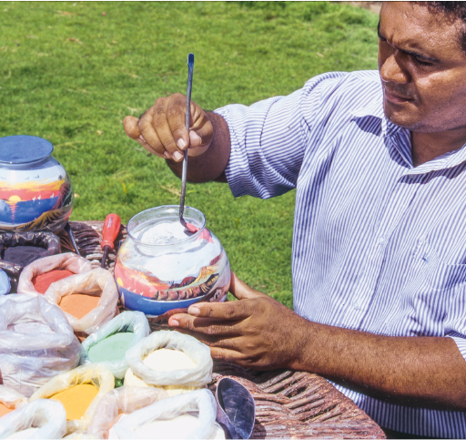 Imagem: Fotografia. Um homem está sentado com a mão esquerda sobre um pote de vidro com areia colorida dentro. Com a mão direita, ele segura um objeto fino e comprido dentro do pote. Na frente dele há vários saquinhos com areia colorida sobre uma mesa.  Fim da imagem.