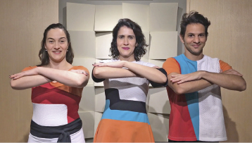 Imagem: Fotografia. Duas mulheres e um homem com camisetas coloridas estão com os braços na frente do corpo. Os braços estão cruzados e as mãos em cima dos cotovelos.    Fim da imagem.