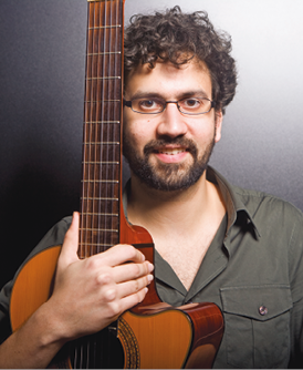 Imagem: Fotografia. Jonas, homem com cabelo encaracolado e curto, óculos, barba e bigode está sorrindo e segurando um violão.  Fim da imagem.