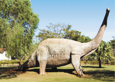 Imagem: Fotografia. Réplica de um dinossauro com pescoço e rabo compridos. Em volta há muitas árvores e ao fundo, uma casa. Fim da imagem.