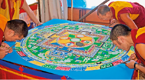 Imagem: Fotografia. Quatro monges com roupa amarela e vermelha estão segurando instrumentos finos e compridos e com os corpos inclinados para frente, sobre uma mesa grande com uma pintura colorida em cima.  Fim da imagem.