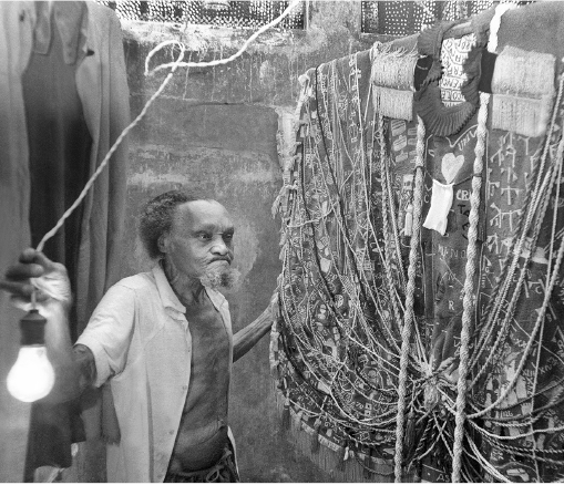 Imagem: Fotografia em preto e branco. Um homem com cabelo encaracolado e barba volumosa está com camisa aberta e segurando uma corda com uma lâmpada acesa na ponta. Na frente dele há uma escultura feita de cordas e tecidos. Fim da imagem.