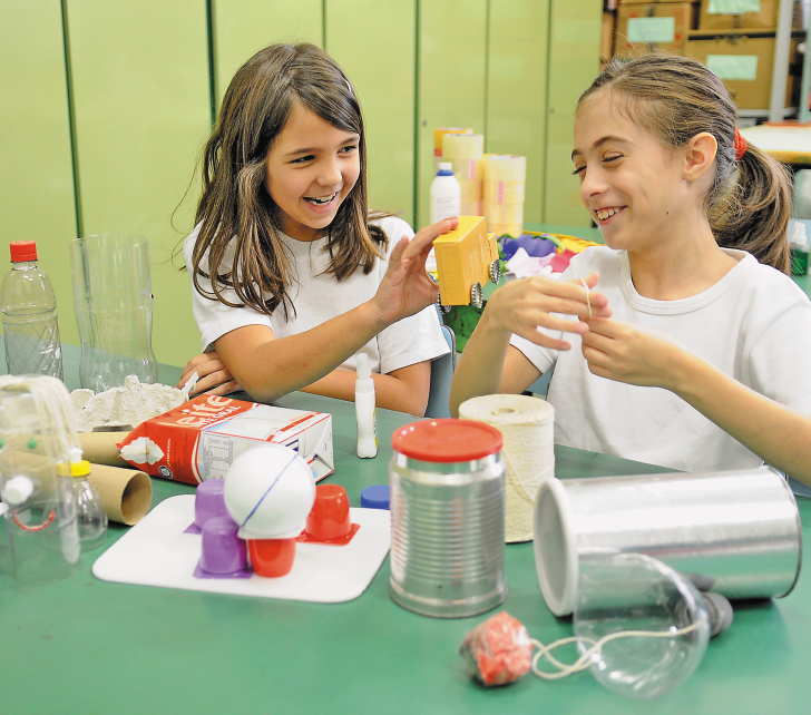 Imagem: Fotografia. Duas meninas com camiseta branca estão sentadas atrás de uma mesa e sorrindo. À esquerda, uma delas está segurando um carrinho feito de papel. À direita, a outra está segurando uma flor colorida de papel. Sobre a mesa há vários objetos. Fim da imagem.