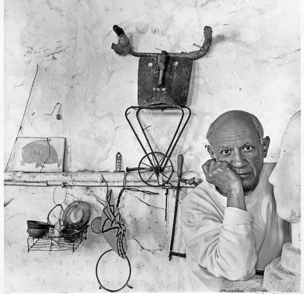 Imagem: Fotografia em preto e branco. À direita, Pablo Picasso, homem calvo com olhos grandes, nariz largo e lábios finos. Ele está com a mão direita apoiada no rosto. À esquerda, esculturas. Fim da imagem.