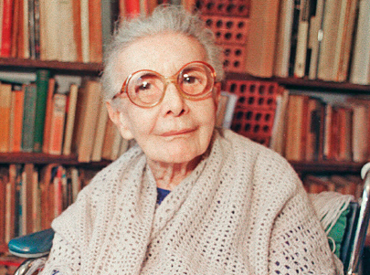 Imagem: Fotografia. Nise, senhora com cabelo grisalho, óculos redondos e lábios finos está sentada e sorrindo. Ao fundo, prateleiras com vários livros. Fim da imagem.