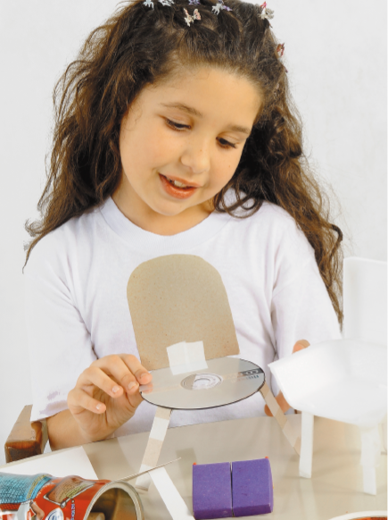 Imagem: Fotografia. Uma menina com presilhas de borboleta na cabeça e camiseta branca está segurando uma cadeira pequena feita com CD e papelão.  Fim da imagem.