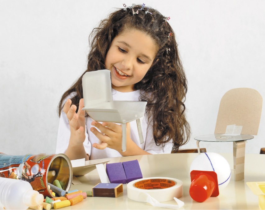 Imagem: Fotografia. A menina está sorrindo e segurando um berço pequeno feito de caixa de isopor. Na frente dela há uma mesa com vários objetos em cima. Fim da imagem.