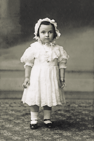 Imagem: Fotografia em preto e branco. Uma criança com chapéu, vestido e sapatos está em pé. Fim da imagem.