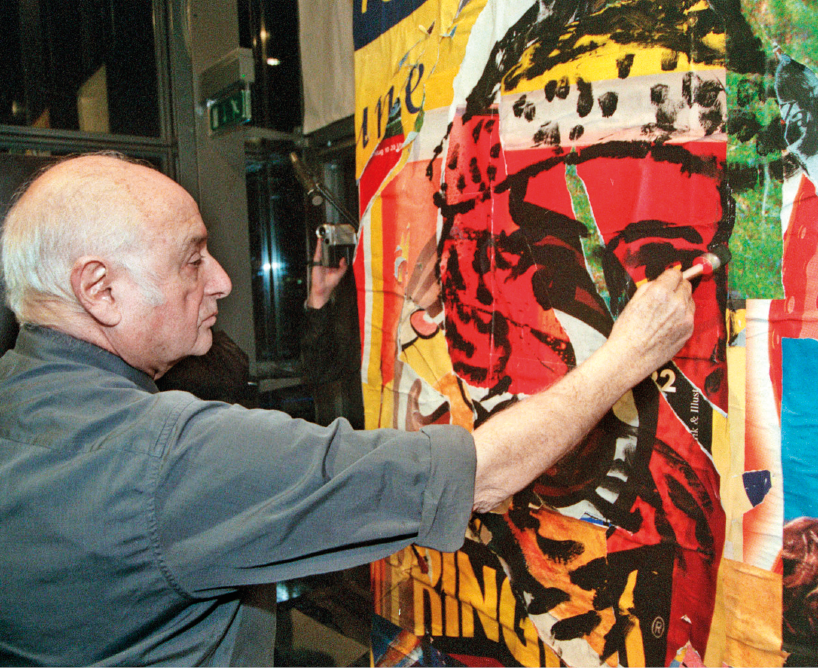 Imagem: Fotografia. Um homem calvo com cabelo grisalho e camisa cinza está sentado e segurando um pincel sobre uma pintura colorida. Fim da imagem.