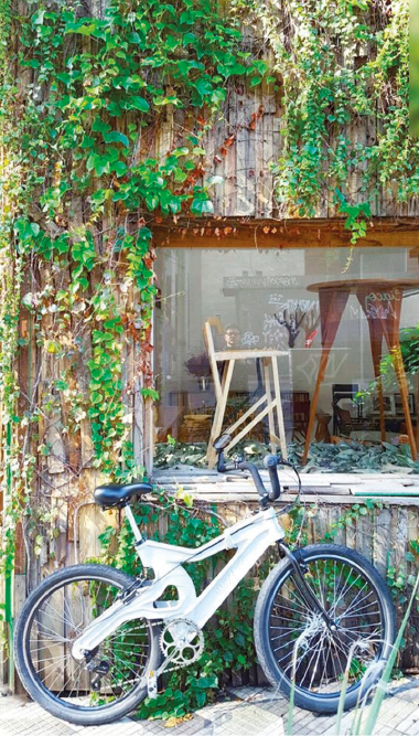 Imagem: Fotografia. Uma bicicleta cinza com rodas pretas. Ao fundo, uma construção de madeira com plantas em volta e no centro há objetos expostos em uma vitrine. Fim da imagem.