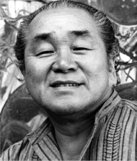 Imagem: Fotografia em preto e branco. Manabu, homem oriental com cabelo penteado para trás, olhos amendoados, nariz largo e lábios finos está sorrindo. Fim da imagem.