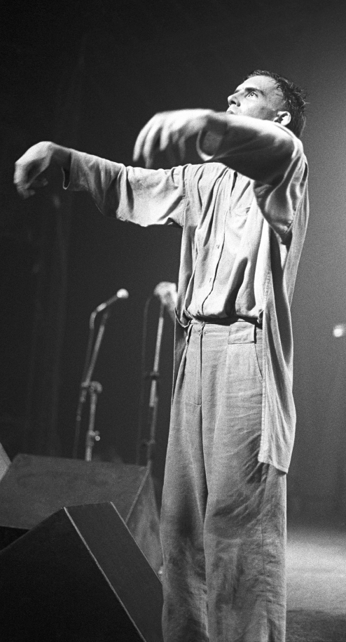 Imagem: Fotografia em preto e branco. Um homem com blusa e calça está em pé com os braços para frente sobre um palco.  Fim da imagem.