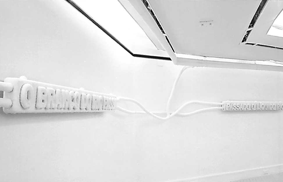 Imagem: Fotografia. Duas placas brancas com palavras em relevo estão conectadas a um teto branco por cabos em uma parede branca. Fim da imagem.