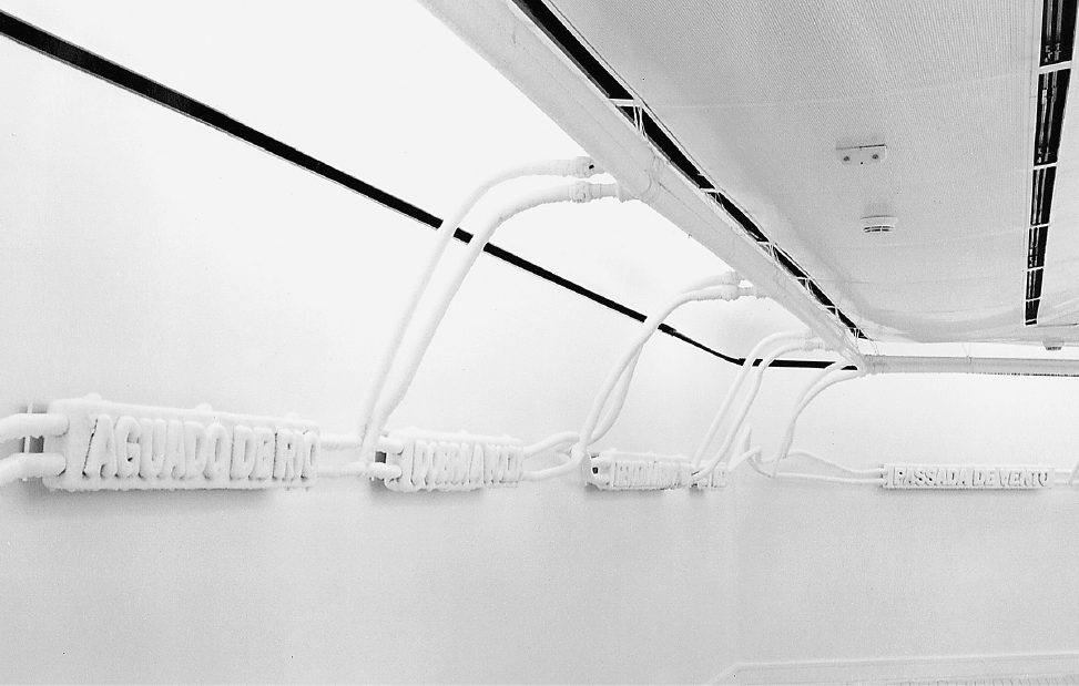 Imagem: Fotografia. Cinco placas brancas com palavras em relevo estão conectadas a um teto branco por cabos em uma parede branca. Fim da imagem.