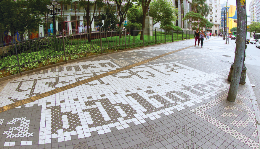 Imagem: Fotografia. Uma calçada com várias palavras em vários idiomas, composto por vários quadrados. Ao fundo, árvores e prédios. Fim da imagem.
