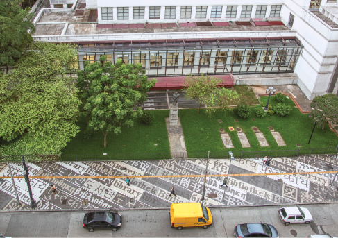 Imagem: Fotografia. Vista aérea de uma calçada em tons de cinza na frente de uma construção grande com árvores em volta.  Fim da imagem.