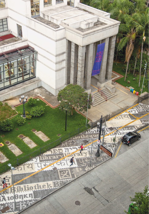 Imagem: Fotografia. Vista aérea do prédio grande e a calçada com palavras em vários idiomas.  Fim da imagem.