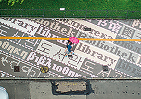 Imagem: Fotografia. Vista de cima da calçada em tons de cinza com palavras em vários idiomas. Fim da imagem.