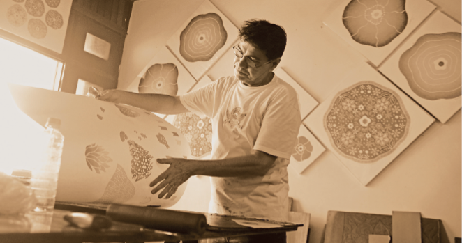 Imagem: Fotografia em sépia. Um homem com cabelo curto, óculos e camiseta está segurando um papel grande com desenhos sobre uma mesa. Ao fundo, quadros pendurados em uma parede. Fim da imagem.