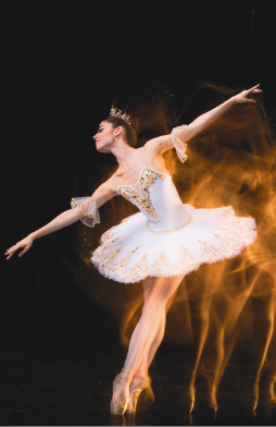 Imagem: Fotografia. Uma mulher com cabelo preso, tiara, vestido e sapatilhas de bailarina está nas pontas dos pés e com os braços abertos. Atrás dela, sombra de seus movimentos. Fim da imagem.