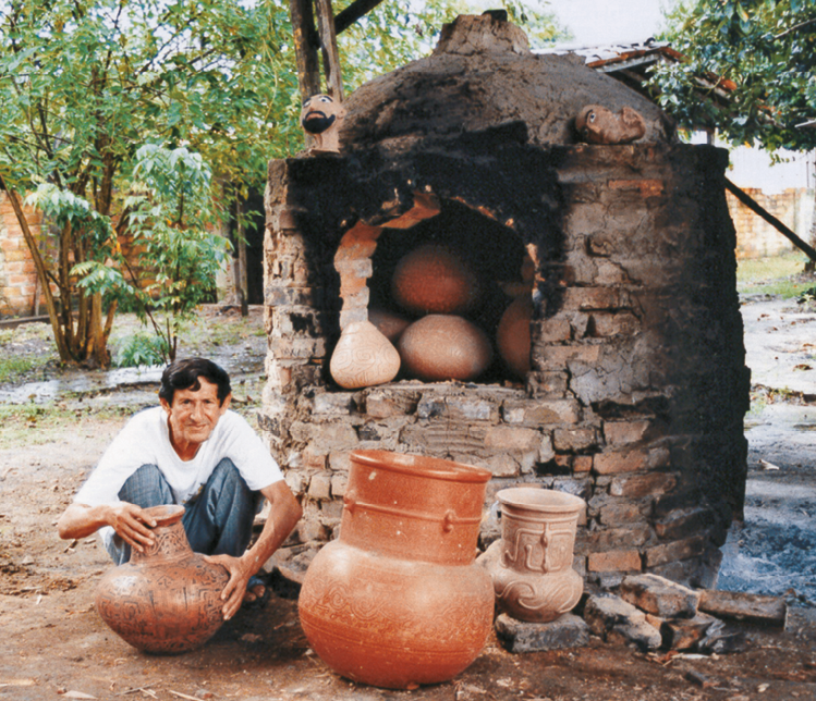 Imagem: Fotografia. Mestre Cardoso está agachado e segurando um vaso de argila no chão. Ao lado há mais vasos e atrás dele, um forno de pedras. Ao fundo, árvores. Fim da imagem.