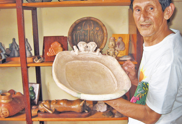 Imagem: Fotografia. Mestre Cardoso, homem com olhos escuros, nariz grande e lábios finos está segurando um recipiente de cerâmica. Atrás dele há vários objetos em uma prateleira.  Fim da imagem.