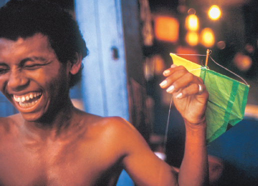 Imagem: Fotografia. Um menino com cabelo encaracolado e curto está sem camisa, sorrindo com os olhos fechados e com a mão esquerda levantada. Ao lado há uma pipa pequena, verde e amarela amarrada na mão. Fim da imagem.