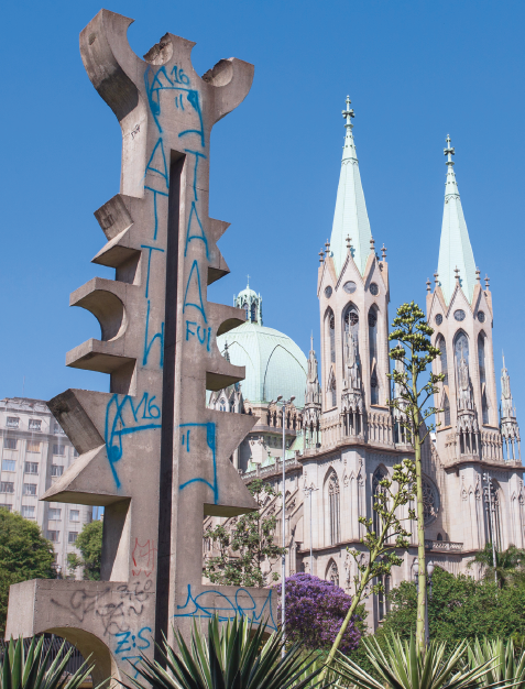 Imagem: Fotografia. Emblema cinza com pichações em volta. Ao fundo, uma catedral grande e plantas. Fim da imagem.