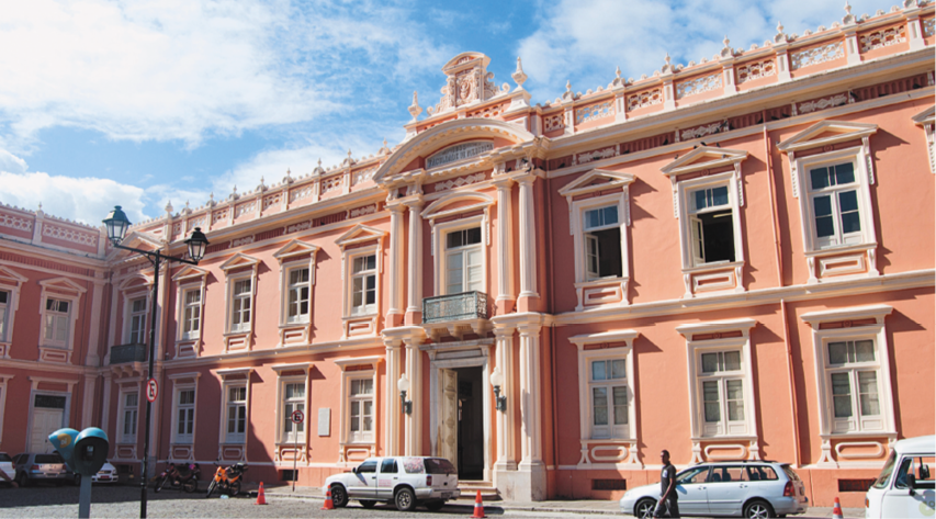 Imagem: Fotografia. Construção grande e extensa com paredes rosa-claro e várias janelas. Fim da imagem.