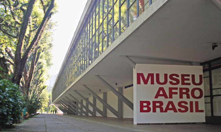 Imagem: Fotografia. Construção grande e extensa. Na parte inferior há uma placa grande com a informação: MUSEU AFRO BRASIL. Ao lado, árvores. Fim da imagem.