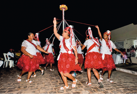 Imagem: Fotografia. Mulheres com camiseta e sandália brancas e saia vermelha estão em roda e segurando fitas com as mãos levantadas. Entre elas há um poste, onde as fitas estão se entrelaçando. Fim da imagem.